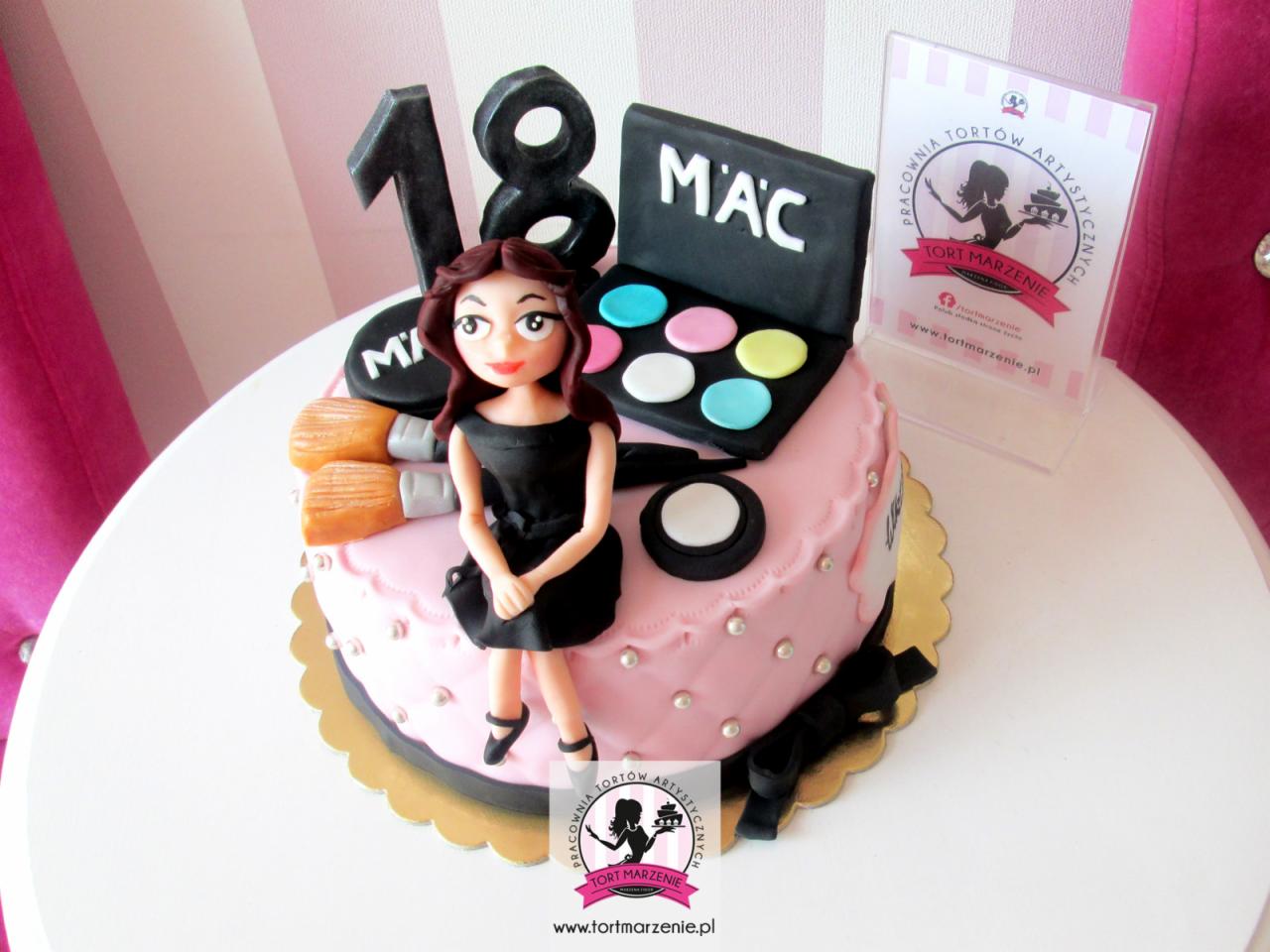 Tort urodzinowy w stylu kosmetycznym z figurką kobiety, paletą cieni i napisem '18', na stole z różowym tłem.
