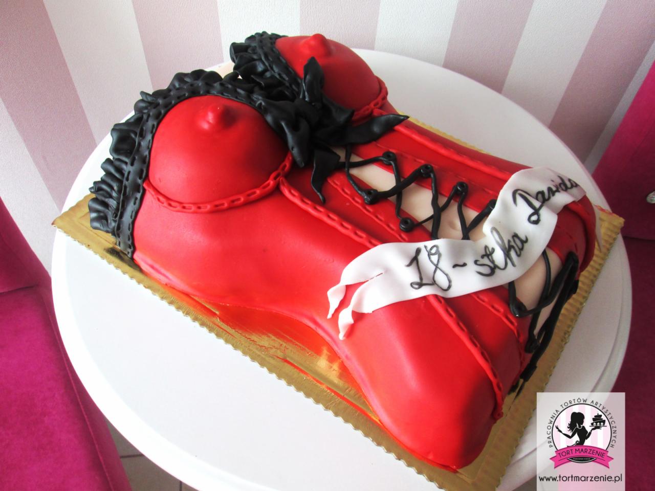 Tort w kształcie czerwonego gorsetu z czarną koronką i sznurowaniem, udekorowany białym napisem '18-stka Dawida', umieszczony na złotej podkładce.