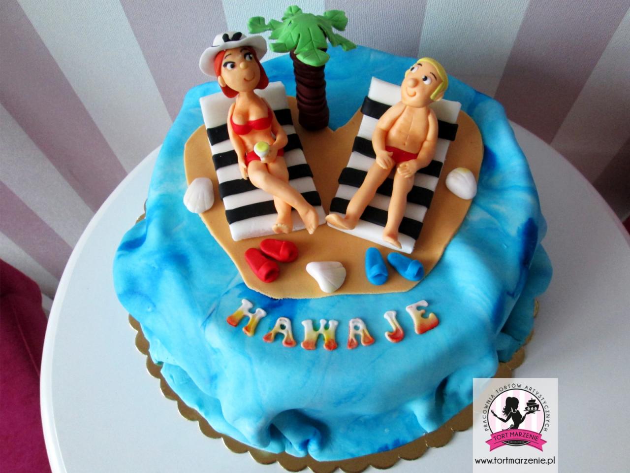 Tort z motywem plażowym: figurki kobiety w bikini i mężczyzny w kąpielówkach na leżakach, palma, muszelki i napis 'HawaJe' na niebieskim tle imitującym morze.