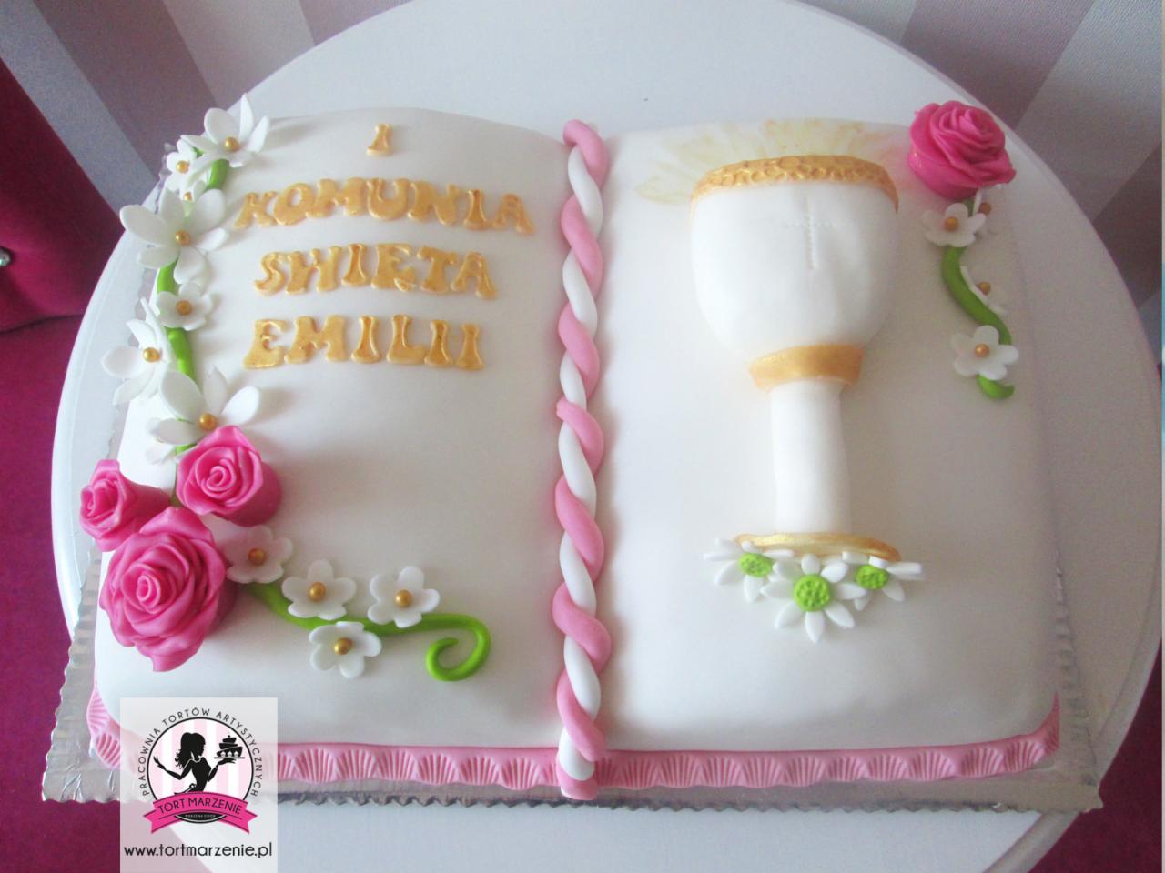 Tort w kształcie otwartej księgi z napisem 'I Komunia Święta Emilii', zdobiony marcepanową hostią, różowymi różami i białymi kwiatkami.