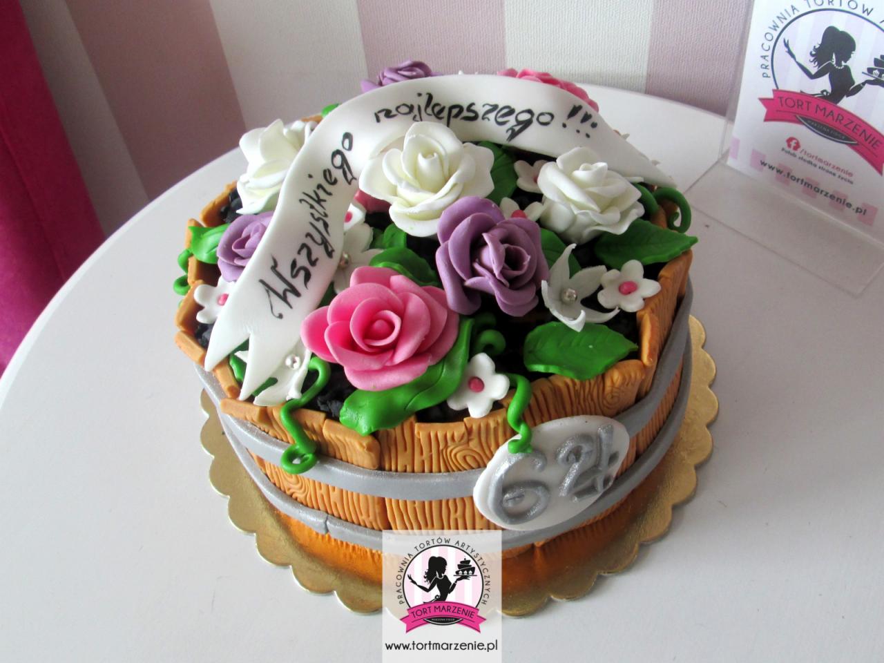 Tort urodzinowy w kształcie drewnianej beczki z kwiatami z lukru i napisem 'Wszystkiego najlepszego!' na białej wstążce, umieszczony na złotym podkładzie.