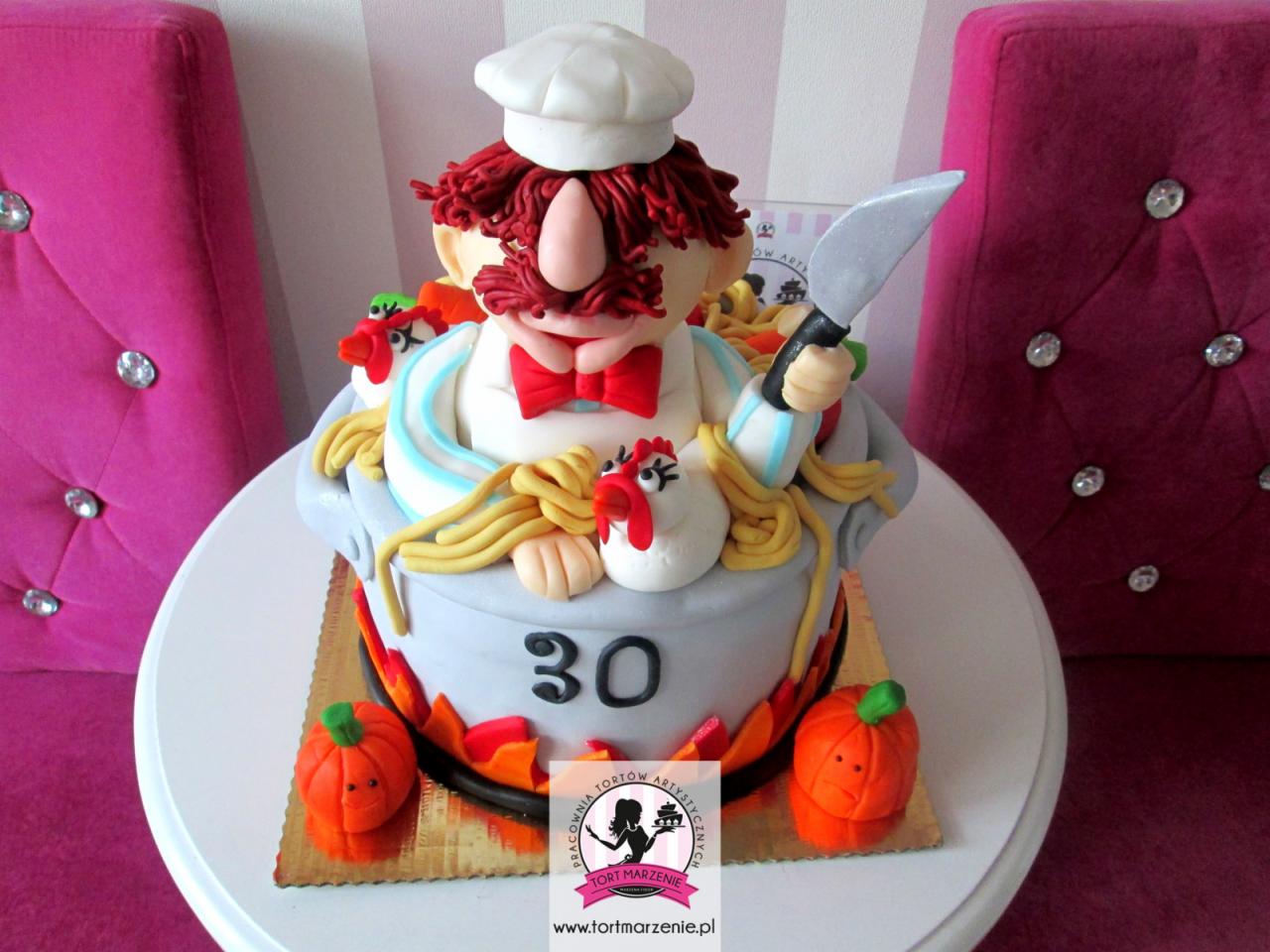 Tort urodzinowy w kształcie garnka z kucharzem, kurczakami i dyniami, ozdobiony cyfrą 30, na białym stoliku z różowymi krzesłami w tle.