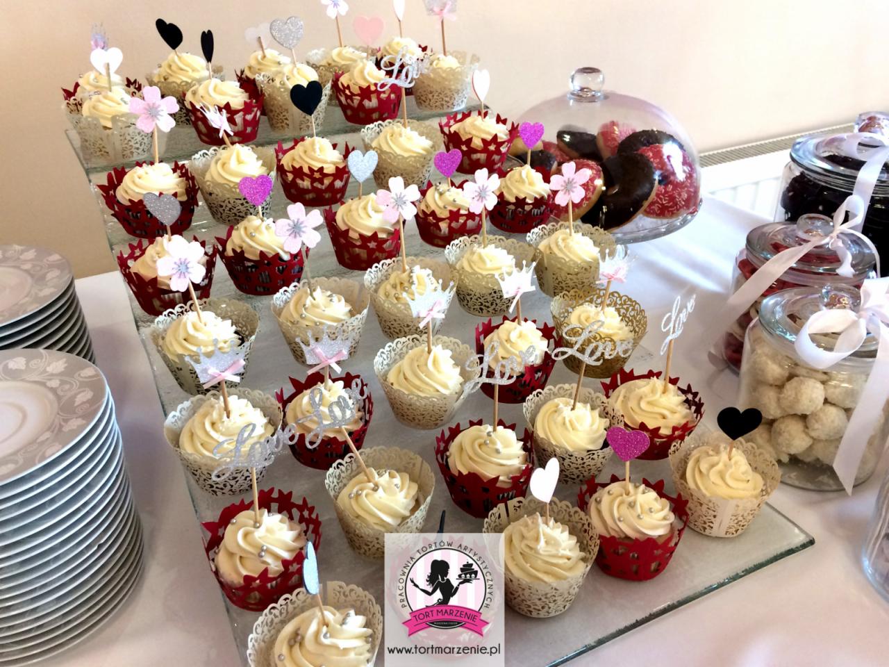 Dekoracyjne babeczki z kremem i topperami na szklanym stojaku, obok słoiki ze słodkościami i stos talerzy