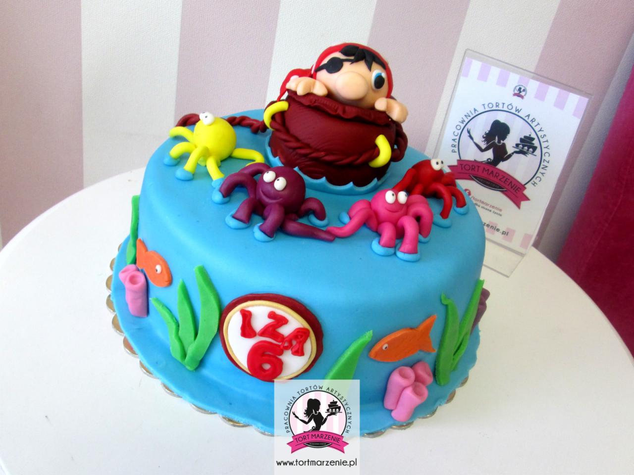 Tort urodzinowy w stylu morskim z figurką pirata wystającą z beczki, otoczony kolorowymi ośmiornicami i dekoracjami z alg i rybek, na froncie tortu napis 'IZA 6'