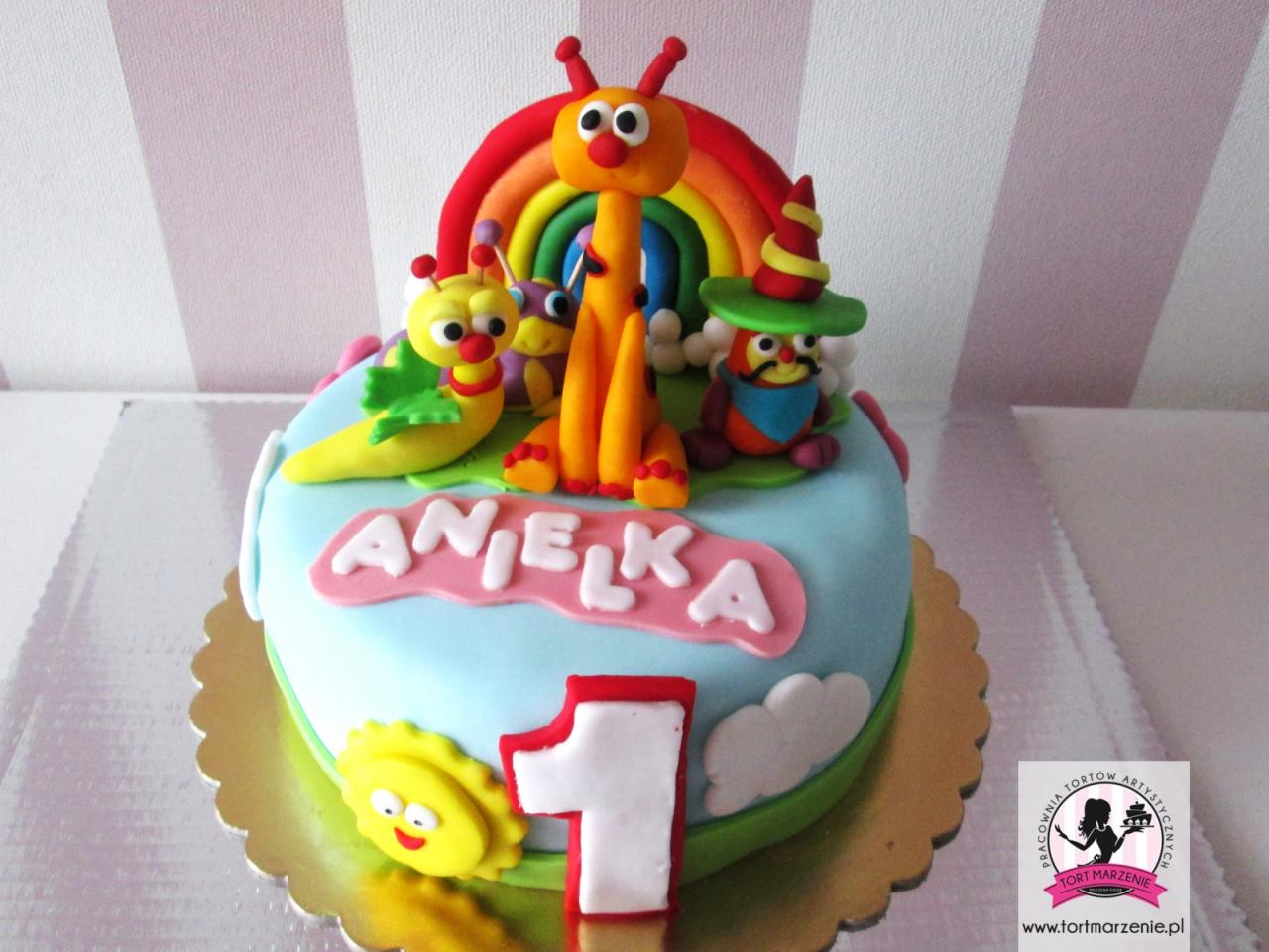 Tort urodzinowy dla dziecka z imieniem Anielka, zdobiony figurkami postaci z bajki i tęczą z lukru, na złotym podkładzie.