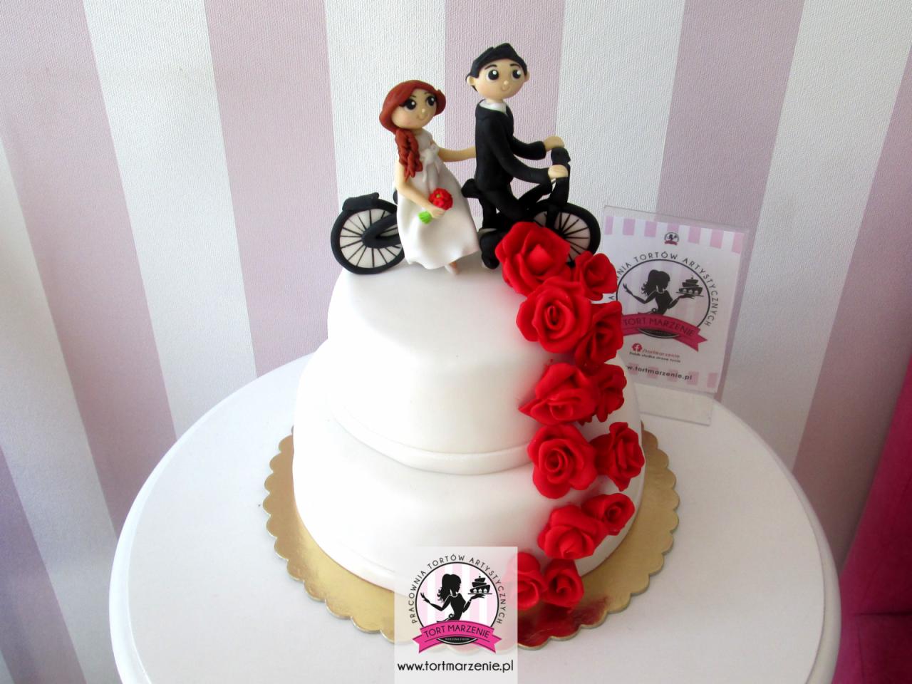 Dwupiętrowy tort weselny z dekoracją z czerwonych róż i figurkami pary młodej na rowerze, umieszczony na złotym podkładzie, z logotypem cukierni w tle.