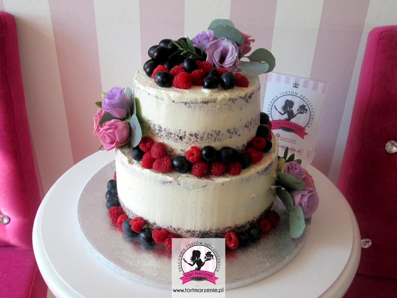 Dwupiętrowy tort 'naked cake' z kremem, udekorowany świeżymi malinami, borówkami, winogronami i kwiatami, na białym stole z różowymi krzesłami w tle, obok widoczny folder reklamowy.