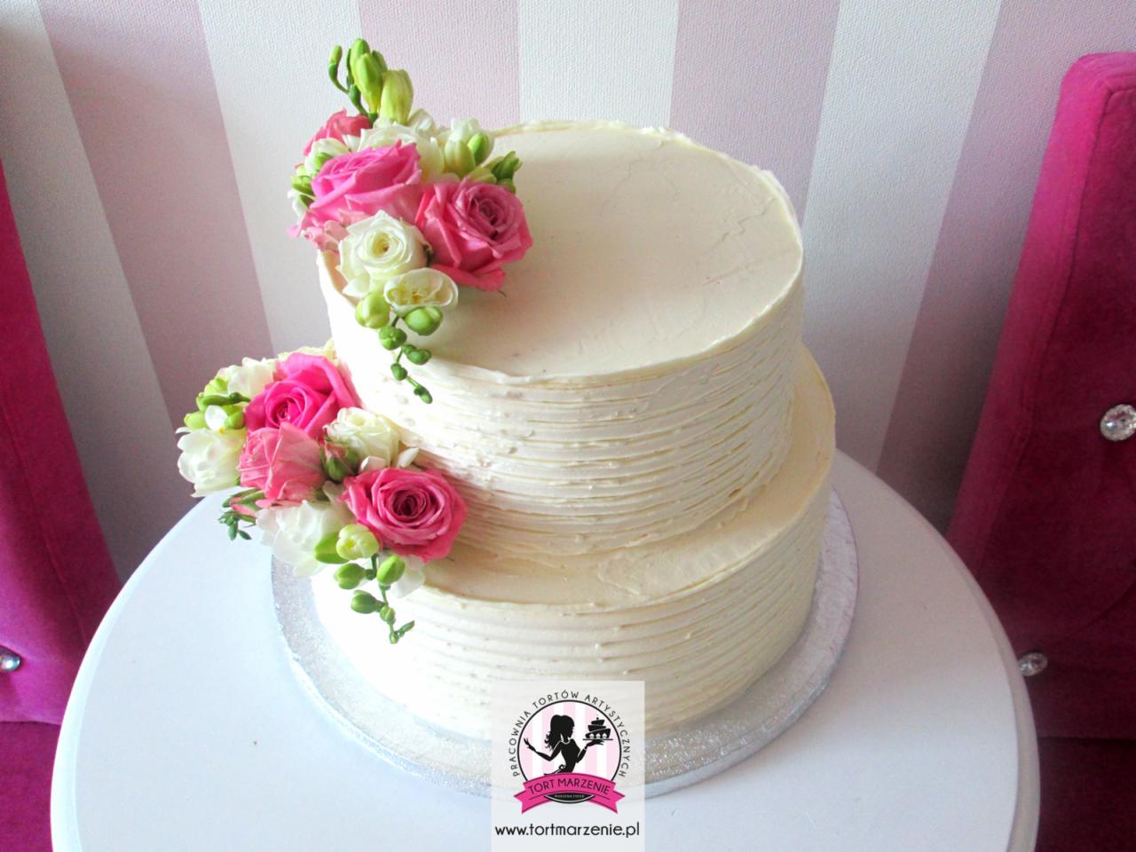 Dwupiętrowy tort weselny z kremowym lukrem i dekoracją z różowych i białych róż oraz frezji, umieszczony na białym stole na tle różowych krzeseł i tapety w paski.