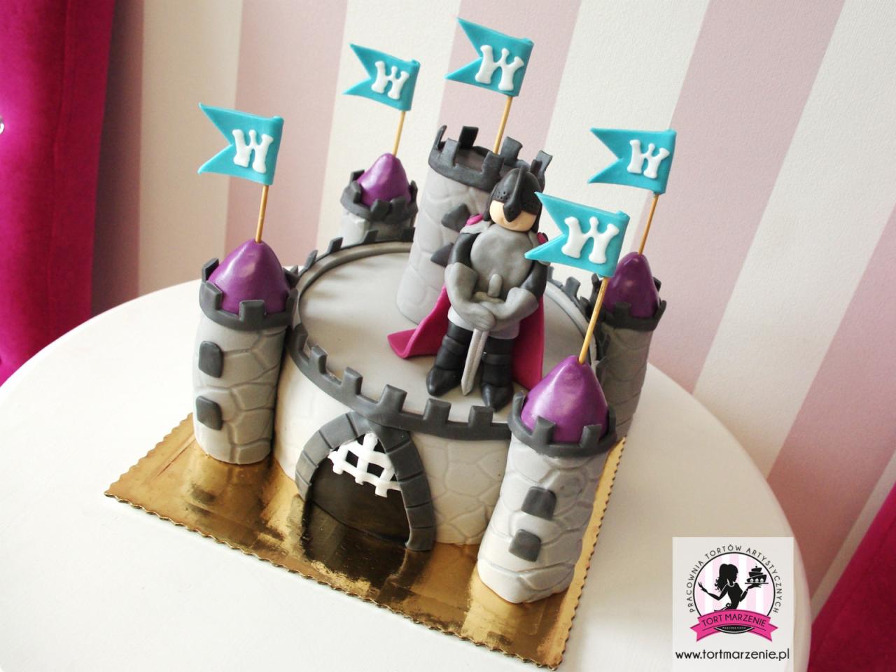 Tort urodzinowy w kształcie zamku z figurką rycerza i flagami z literą 'W' na złotym podkładzie, ustawiony na białym stole z różowo-białym tłem w paski.