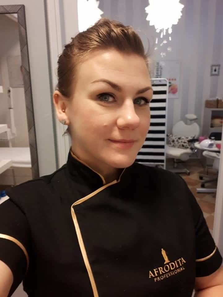 Portret kobiety w czarnym uniformie z logo Afrodita Professional, w tle wyposażenie salonu kosmetycznego i lustro.