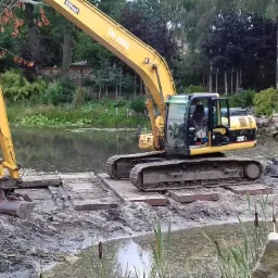 Żółta koparka CAT 320CL stoi na płytach betonowych w płytkim stawie, przygotowując się do pogłębiania, na drugim planie drzewa.