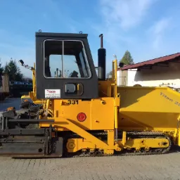 Żółty rozkładacz asfaltu Titan 280 z czarną kabiną i numerem 331, stojący na betonowym placu w słoneczny dzień, z widocznym logo firmy Koliber i innymi pojazdami w tle.