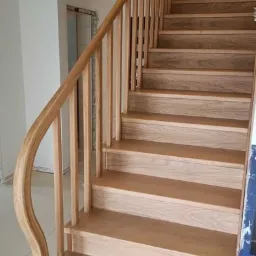 Jasne, drewniane schody z zaokrąglonym poręczą, widok od dołu, w tle surowa ściana i niedokończone wykończenie.