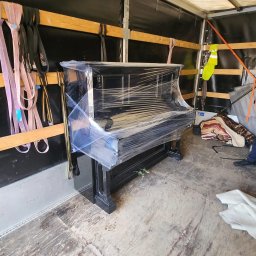 Pan Przeprowadzka - Przeprowadzki Szczecin - Czarny fortepian owinięty folią stretch, zabezpieczony wewnątrz przestrzeni ładunkowej pojazdu, widoczne pasy mocujące i odblaskowa kamizelka.