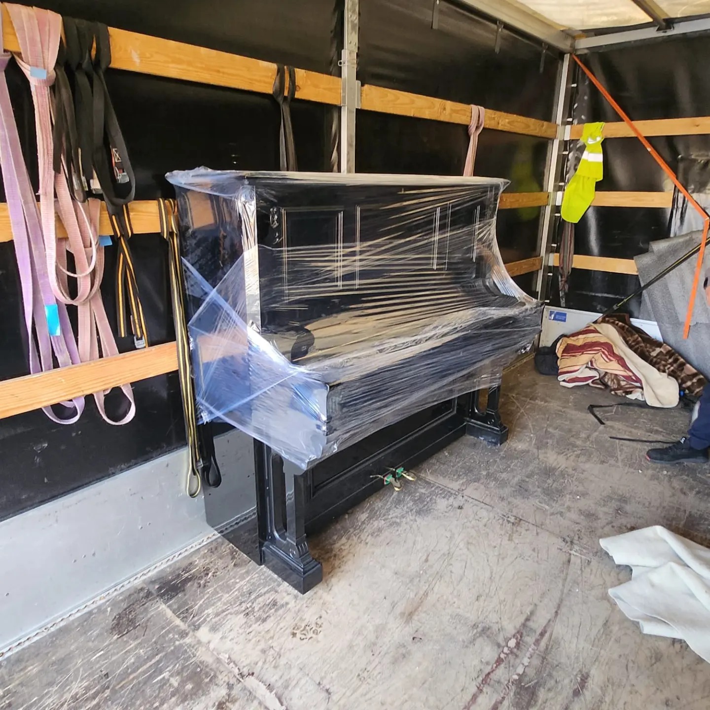 Czarny fortepian owinięty folią stretch, zabezpieczony wewnątrz przestrzeni ładunkowej pojazdu, widoczne pasy mocujące i odblaskowa kamizelka.