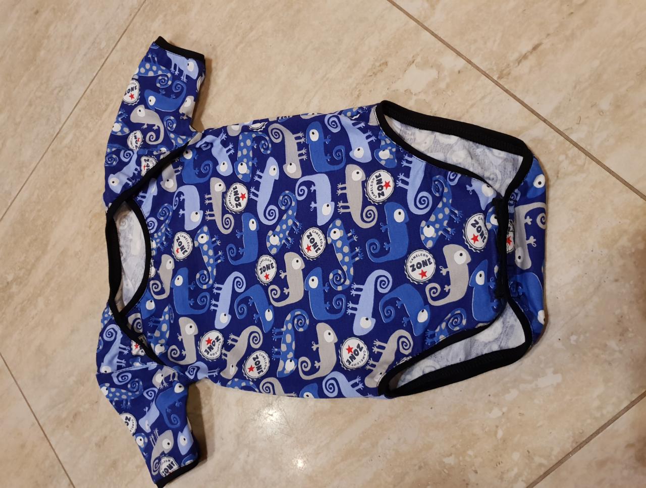 Niebieskie body dziecięce z motywem kameleonów i napisem 'Chameleon Zone', leżące płasko na kafelkach podłogowych.