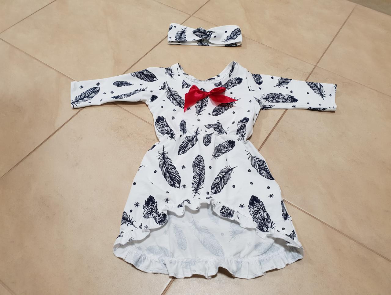 Biała sukienka dla dziewczynki z wzorem piór i czerwoną kokardką, dopasowana opaska na głowę z tym samym wzorem piór, leżąca na kafelkach podłogowych.