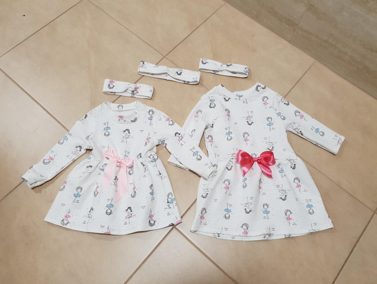 Dwie identyczne sukienki dziecięce z długim rękawem, w biały wzór baletnic, ozdobione różowymi kokardami, leżące na kafelkach podłogowych, wraz z dopasowanymi opaskami na głowę.