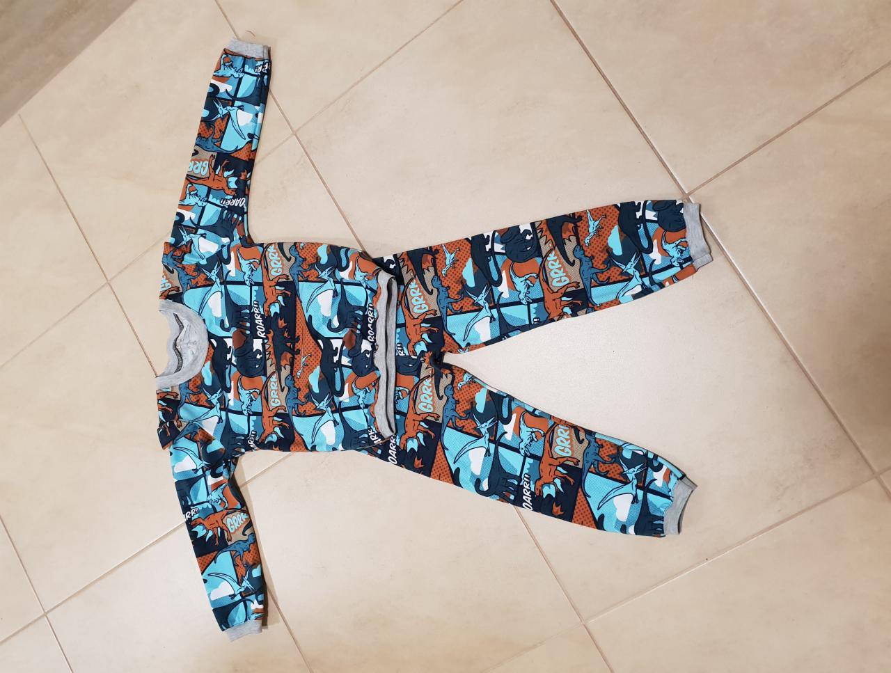 Rozłożona piżama dziecięca z długim rękawem i nogawkami, w niebiesko-brązowe dinozaury na białym tle płytek podłogowych.