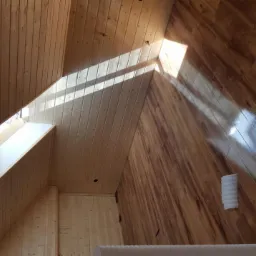 Wnętrze poddasza z drewnianą boazerią na ścianach i suficie, z oknem dachowym wpuszczającym snop światła, oraz połyskującą drewnianą podłogą.