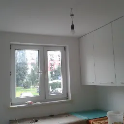 Wnętrze kuchni w trakcie remontu: świeżo pomalowane ściany na biało, okno z widokiem na zieleń, białe szafki kuchenne pod sufitem, goła żarówka zwisająca z sufitu, zabezpieczone blaty.