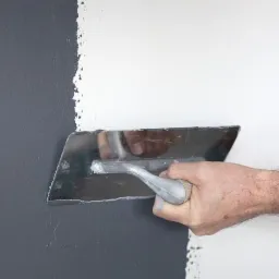 Mężczyzna rozprowadza masę szpachlową na ścianie, widoczna granica między surową szarą ścianą a świeżo nałożoną białą warstwą, używając metalowej szpachelki.