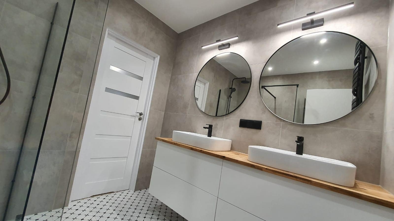 Nowoczesna łazienka w szarościach z dwoma okrągłymi lustrami, białymi umywalkami nablatowymi na drewnianym blacie i wzorzystą podłogą.