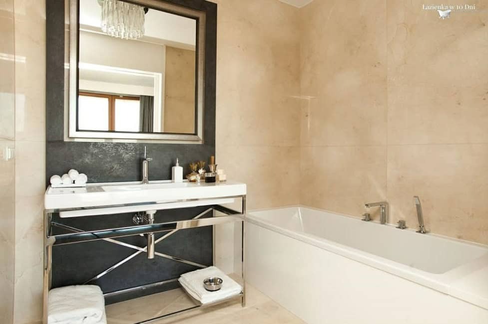 Elegancka łazienka z beżowymi płytkami, białą wanną i umywalką na chromowanym stelażu. Nad umywalką lustro w ciemnej ramie i dekoracyjny żyrandol.