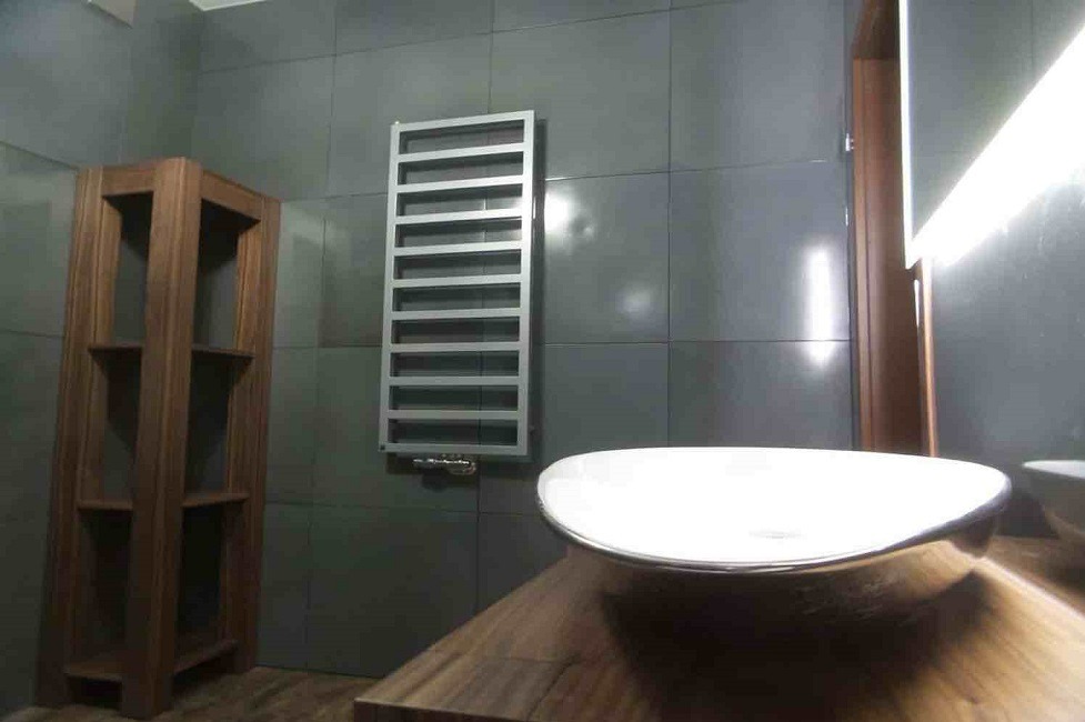Nowoczesna łazienka z szarymi płytkami, drewnianą szafką i grzejnikiem drabinkowym. Umywalka nablatowa o owalnym kształcie. Minimalistyczny design.