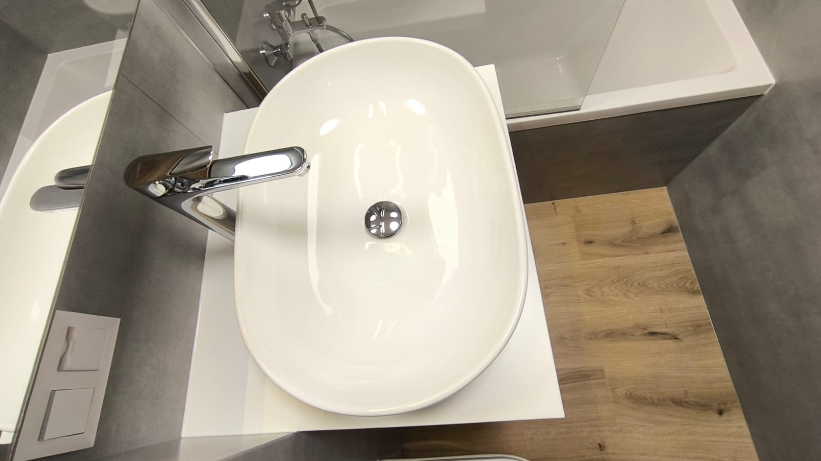 Biała, owalna umywalka z chromowaną baterią, zamontowana na białym blacie. W tle widoczna wanna z parawanem i szare płytki ścienne oraz podłoga imitująca drewno.