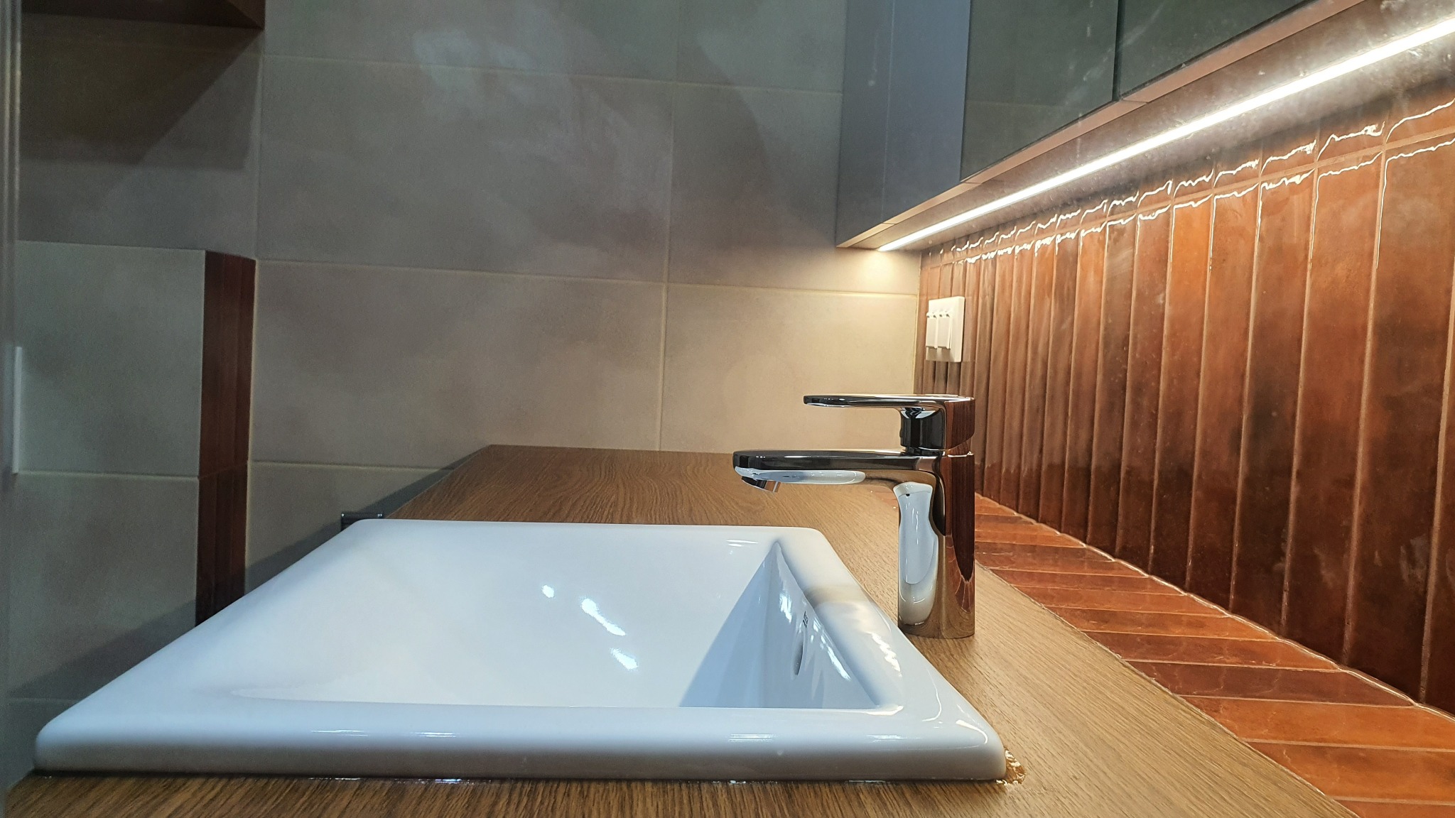 Biała, prostokątna umywalka z chromowaną baterią na blacie z drewnopodobnego materiału. W tle beżowe płytki i pionowe, brązowe płytki podświetlone listwą LED.