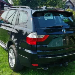 Błyszczący, czarny SUV BMW X3 zaparkowany na trawie, widok z tyłu, po korekcie lakieru, z widocznym odbiciem nieba i drzew w karoserii.