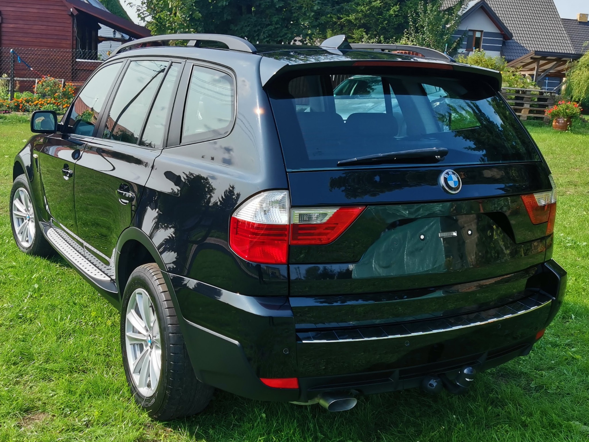 Błyszczący, czarny SUV BMW X3 zaparkowany na trawie, widok z tyłu, po korekcie lakieru, z widocznym odbiciem nieba i drzew w karoserii.