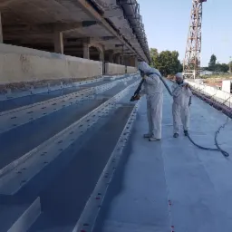 Pracownicy w kombinezonach ochronnych aplikują izolację na betonowe trybuny stadionu z widoczną konstrukcją dźwigu w tle.