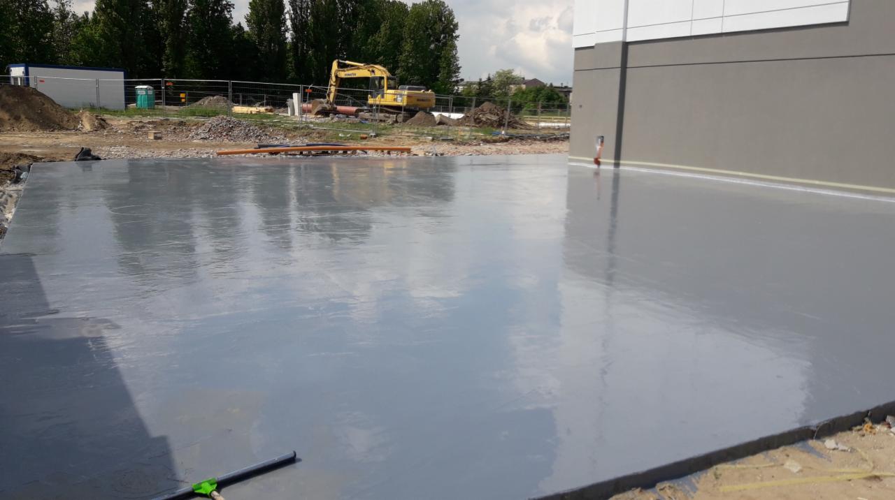 Świeżo wylana, gładka, szara posadzka przemysłowa na placu budowy, z widocznym w tle sprzętem budowlanym i fragmentem budynku.