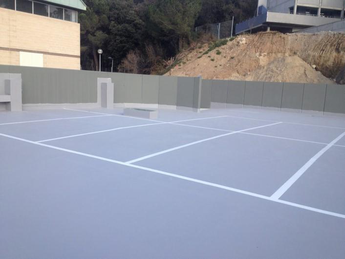 Szara powierzchnia dachu podzielona białymi liniami na kwadraty, otoczona szarym murem, z widocznym budynkiem i zboczem wzgórza w tle.