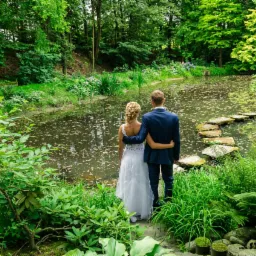 Para młoda w objęciach stoi nad stawem w otoczeniu zieleni, kamienne stopnie prowadzą przez wodę, widok z tyłu.