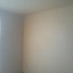 Wnętrze pokoju po nałożeniu jasnej, gładkiej powłoki na ściany, widoczne okno z częściowo zamaskowaną szybą i fragment ciemnej framugi drzwi.