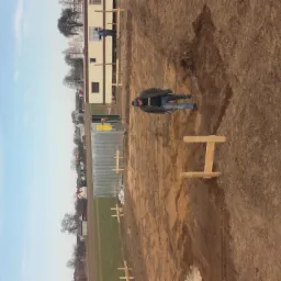 Pracownik w wykopie pod fundamenty, widoczne drewniane szalunki, teren przygotowany pod wylewkę, barak budowlany w tle.