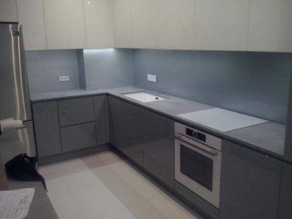 Szara kuchnia z błyszczącymi frontami szafek, szklany panel nad blatem, wbudowany piekarnik i minimalistyczny zlew.