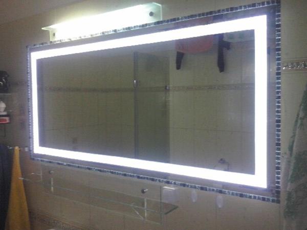 Prostokątne lustro z podświetleniem LED i mozaikową ramą w łazience z beżowymi kafelkami, widoczne szklane półki pod lustrem.