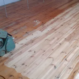 Drewniana podłoga częściowo wyszlifowana szlifierką marki Hummel, widoczny kontrast między surowym, jasnym drewnem a fragmentem po obróbce, z widocznymi śladami starego lakieru.