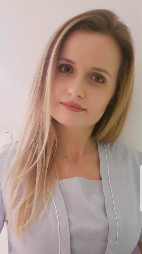 Portret kobiety o blond włosach w pastelowej bluzce, z delikatnym makijażem i subtelną biżuterią na szyi, patrzącej wprost w obiektyw.