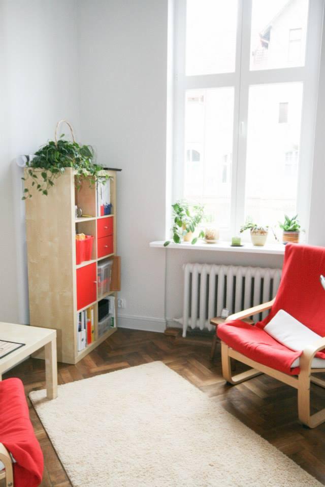 Jasne wnętrze gabinetu z czerwonym fotelem, regałem wypełnionym książkami i roślinami doniczkowymi na parapecie okna.