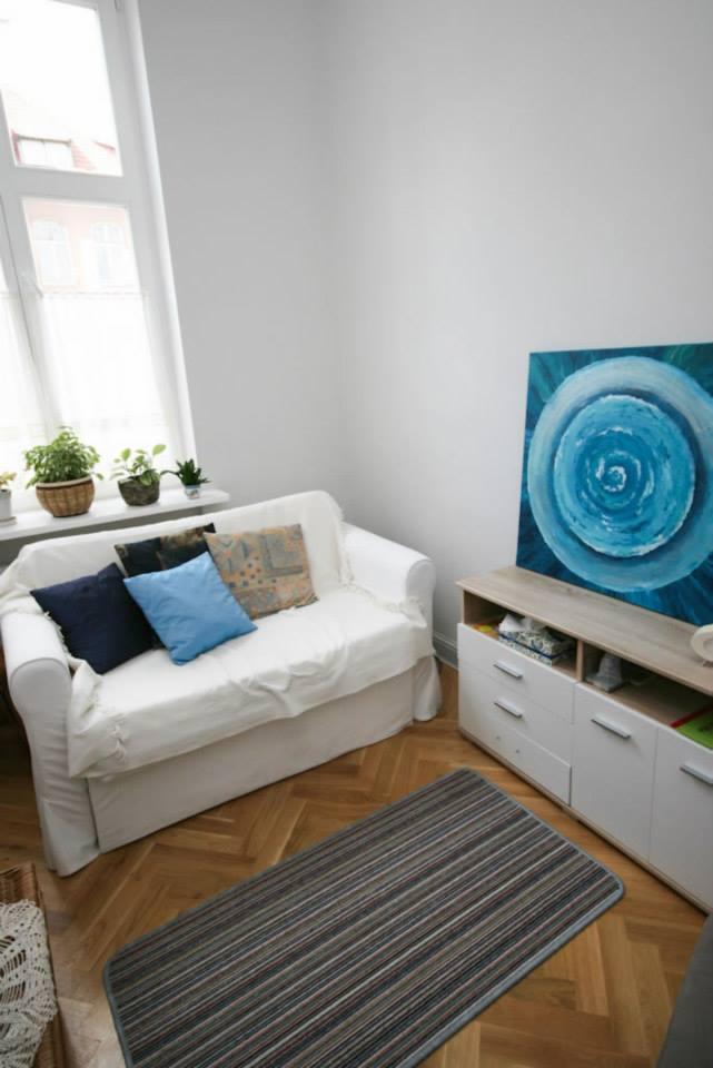 Jasny pokój z białą sofą z poduszkami, obrazem z motywem spirali na komodzie i dywanem w paski na parkiecie.