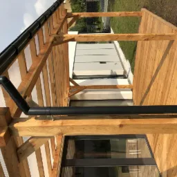 Drewniana konstrukcja zadaszenia tarasu z widocznym systemem odprowadzania wody i fragmentem elewacji budynku.