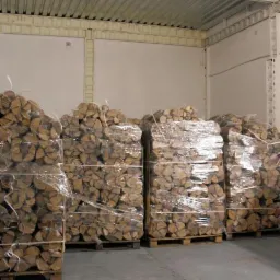 Palety z pociętym, suszonym drewnem dębowym kominkowym, zapakowane w folię termokurczliwą, składowane w magazynie z betonową podłogą i metalowym sufitem.