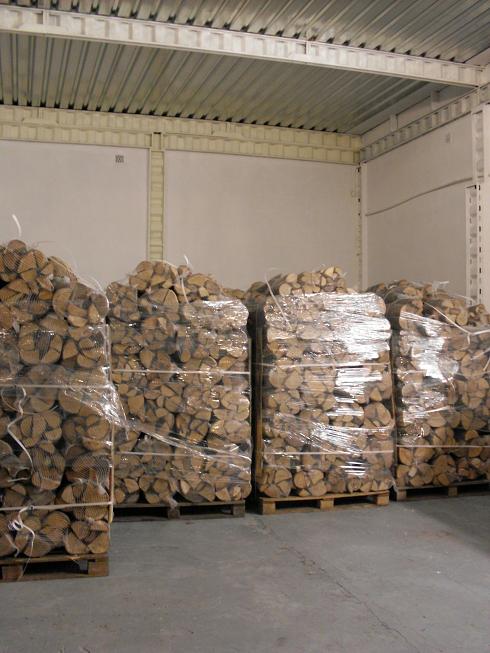 Palety z pociętym, suszonym drewnem dębowym kominkowym, zapakowane w folię termokurczliwą, składowane w magazynie z betonową podłogą i metalowym sufitem.