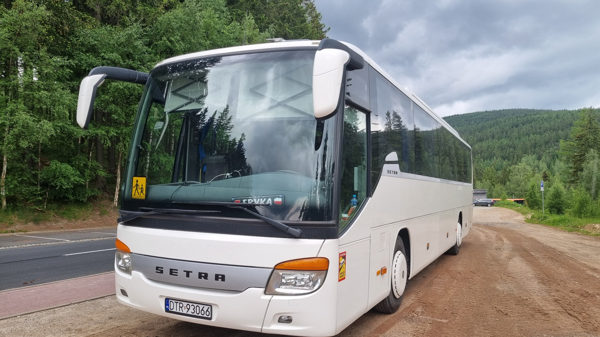 Biały autokar Setra zaparkowany na żwirowej drodze, w tle zalesione wzgórze i pochmurne niebo. Na szybie naklejka z napisem 'Ervuka' i flaga Polski, a także żółty znak ostrzegawczy z sylwetkami...