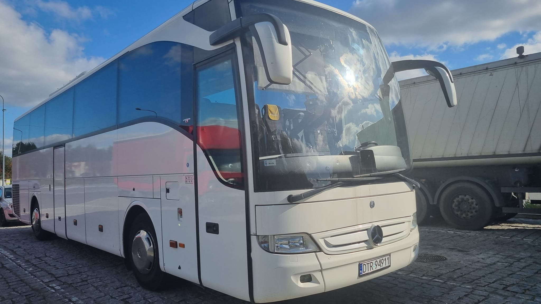 Biały autokar Mercedes z widoczną tablicą rejestracyjną DTR 94911, zaparkowany na brukowanej nawierzchni, z odbiciem chmur w szybach.